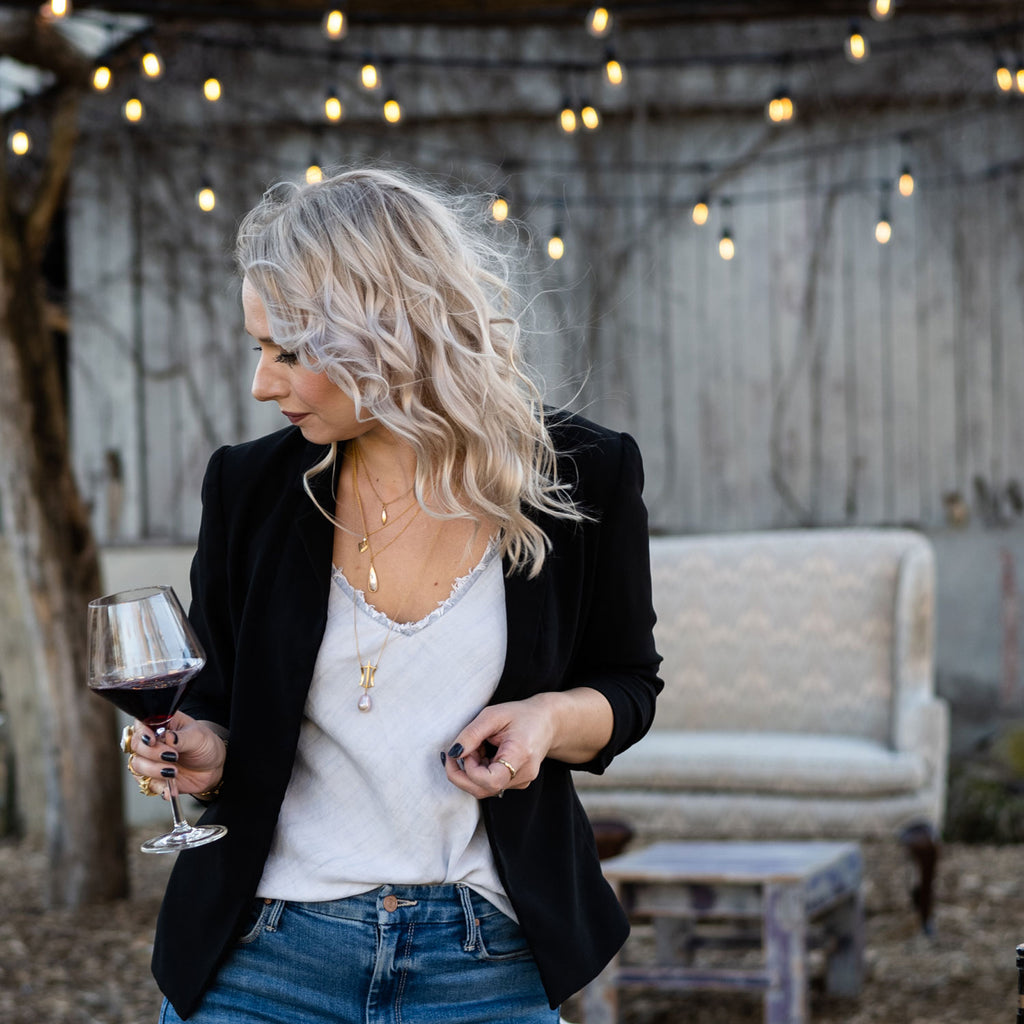 woman holding glass of wine while wearing layers of gold, diamond, and pink pearl necklaces from Nikki Lorenz Designs