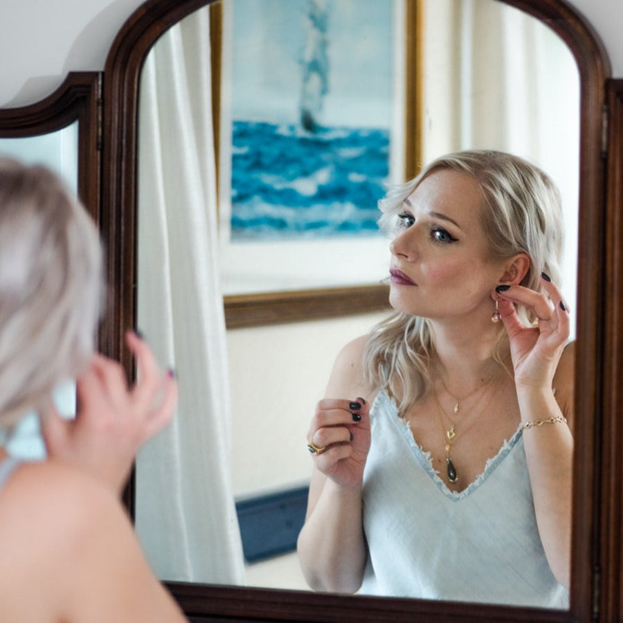 woman wearing layered necklaces looking in mirror while putting on lavender pearl and zircon earrings from Nikki Lorenz Designs
