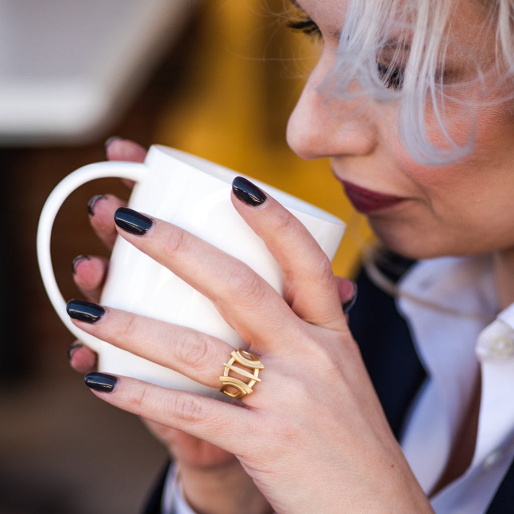 woman with dark finger nail polish holding a coffee mug while wearing bold, modern gold ring from Nikki Lorenz Designs